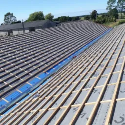 Konstrukcja dachu z widocznymi łatami, membraną dachową i niebieską folią wzdłuż kalenicy, w tle budynki gospodarcze i zielony krajobraz.