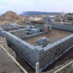 Wznoszone ściany fundamentowe z bloczków betonowych na budowie domu jednorodzinnego, widoczny stos bloczków, poziomica i pracownik w oddali.