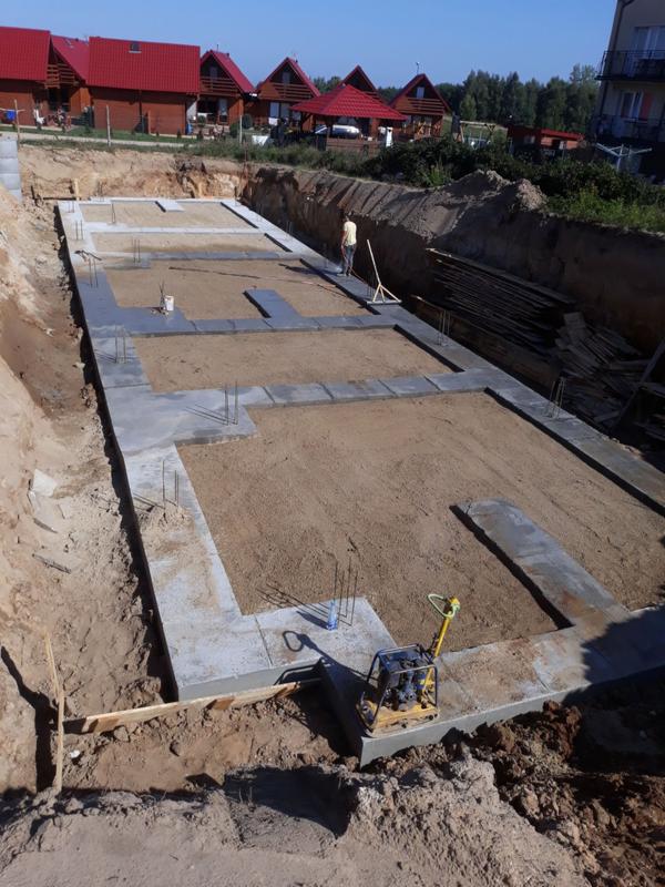 Przygotowanie terenu pod budowę domu jednorodzinnego: wykopane fundamenty z częściowo wylanym betonem, widoczna zagęszczarka i pracownik z grabiami w tle, domy w tle.