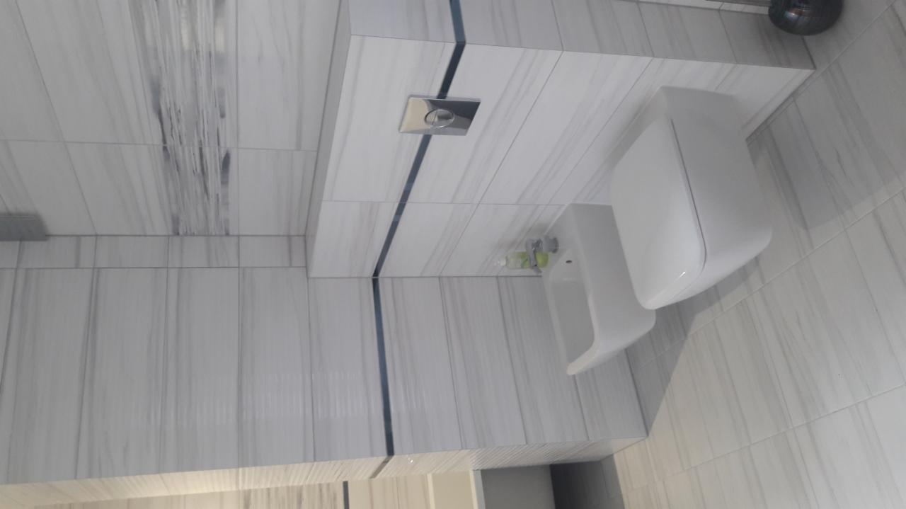 Nowoczesna toaleta podwieszana z białą ceramiką, zamontowana na ścianie wyłożonej płytkami imitującymi marmur z ciemnymi fugami, widoczny przycisk spłuczki i dozownik mydła.