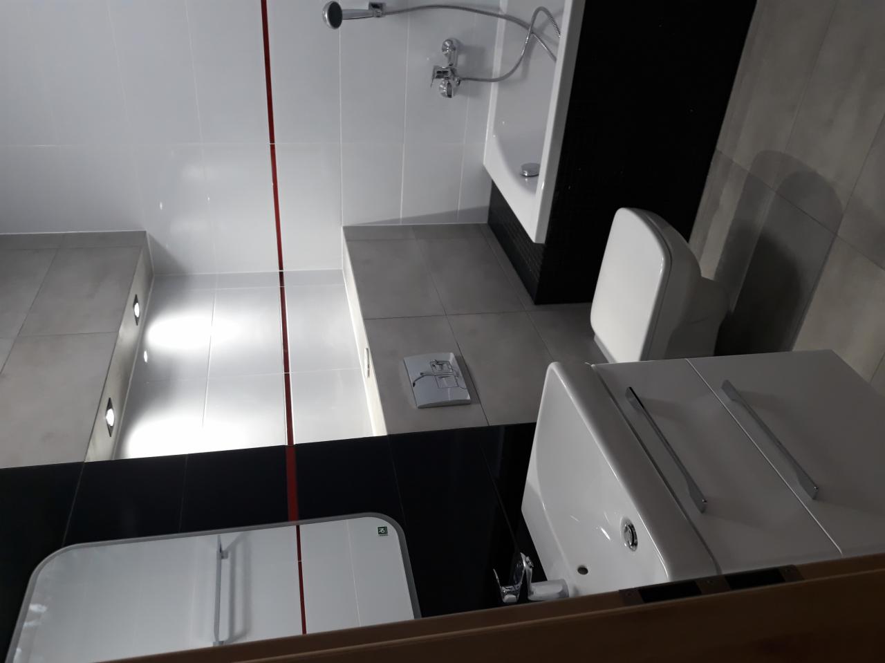 Nowoczesna łazienka z białymi płytkami, czarnymi akcentami i elementami armatury, widok na umywalkę z szafką, toaletę i fragment wanny z prysznicem.