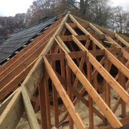 Herba-Tech - Konstrukcja drewniana dachu w trakcie budowy, widoczne krokwie, izolacja z wełny mineralnej i fragment pokrycia dachowego z szarej dachówki, drzewa w tle.