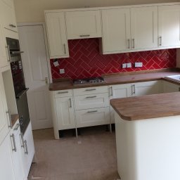 Herba-Tech - Kremowa kuchnia z czerwonymi płytkami ułożonymi w jodełkę, drewnianym blatem i wyspą kuchenną, na podłodze widoczne zabezpieczenia przed uszkodzeniem.