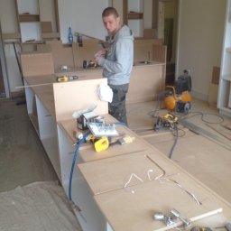 Herba-Tech - Mężczyzna w szarej bluzie roboczej montuje meble z płyty wiórowej w pomieszczeniu z narzędziami, w tle kompresor i piła tarczowa.