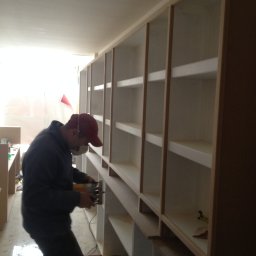 Herba-Tech - Pracownik w masce szlifuje drewniane elementy konstrukcji szafy wnękowej z widocznymi półkami w trakcie montażu.