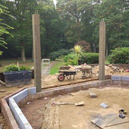 Herba-Tech - Fundamenty pod budowę tarasu z dwoma betonowymi słupami, widoczne zbrojenia, bloczki betonowe, dwie taczki i narzędzia w ogrodzie.