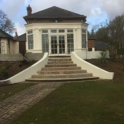 Neoklasycystyczna oranżeria z białymi ramami okiennymi i przeszklonymi drzwiami, schody z kostki brukowej prowadzące do wejścia, świeżo ułożona ścieżka z kostki, trawnik i nasadzenia wokół.