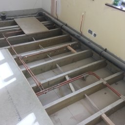 Herba-Tech - Podłoga w trakcie remontu: drewniana konstrukcja, rury miedziane, przewody elektryczne, częściowo położone płyty podłogowe, widoczne okno i ściana z instalacją elektryczną.