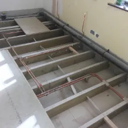 Podłoga w trakcie remontu: drewniana konstrukcja, rury miedziane, przewody elektryczne, częściowo położone płyty podłogowe, widoczne okno i ściana z instalacją elektryczną.