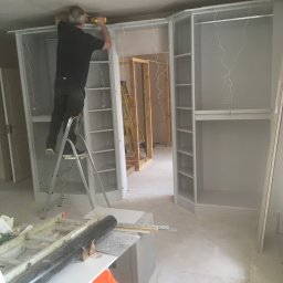 Herba-Tech - Montaż szafy wnękowej: mężczyzna na drabinie w trakcie mocowania górnej listwy szafy za pomocą wkrętarki, widoczne półki i drążki, surowy stan wykończenia wnętrza z widoczną instalacją elektryczną...