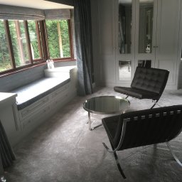 Herba-Tech - Szara szafa wnękowa z lustrami, wbudowane siedzisko przy oknie z szufladami, okrągły stolik kawowy i dwa skórzane fotele Barcelona na miękkim dywanie.