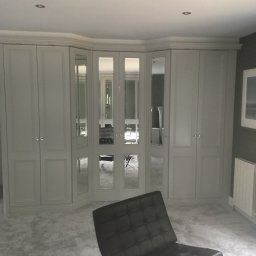 Herba-Tech - Jasnoszara szafa wnękowa z lustrzanymi drzwiami, narożna, z widocznym odbiciem wnętrza i czarnym fotelem Barcelona na tle jasnego dywanu.