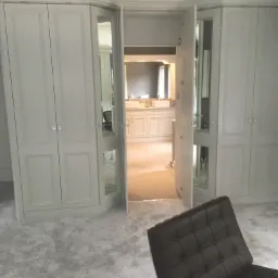 Szara szafa wnękowa z lustrami, otwarte drzwi prowadzą do łazienki z białymi szafkami, widoczny fragment brązowego fotela na pierwszym planie.