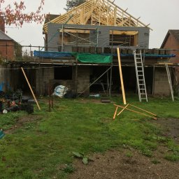 Herba-Tech - Budowa domu w trakcie realizacji: widoczne rusztowania, drewniana konstrukcja dachu, bloczki betonowe, materiały budowlane i drabina na tle zielonego trawnika.