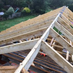 Herba-Tech - Konstrukcja drewniana dachu w trakcie budowy, widoczne krokwie, łaty, poziomice i młotek ciesielski, w tle ogród z placem zabaw.