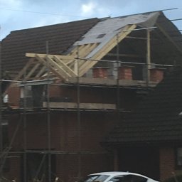 Herba-Tech - Budowa poddasza z drewnianą konstrukcją dachu i ceglanymi ścianami szczytowymi, zabezpieczona folią, widoczna z rusztowania na tle domu z brązową dachówką i białym samochodem.