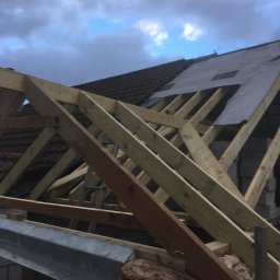 Herba-Tech - Konstrukcja drewniana dachu w trakcie budowy, widoczne krokwie, łaty, fragment pokrycia dachowego i izolacja, pochmurne niebo w tle.