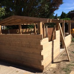 Herba-Tech - Konstrukcja z jasnego drewna w trakcie budowy, prawdopodobnie altana lub domek narzędziowy, z widocznymi elementami szkieletu i deskami tworzącymi ściany, na tle zieleni i błękitnego nieba.