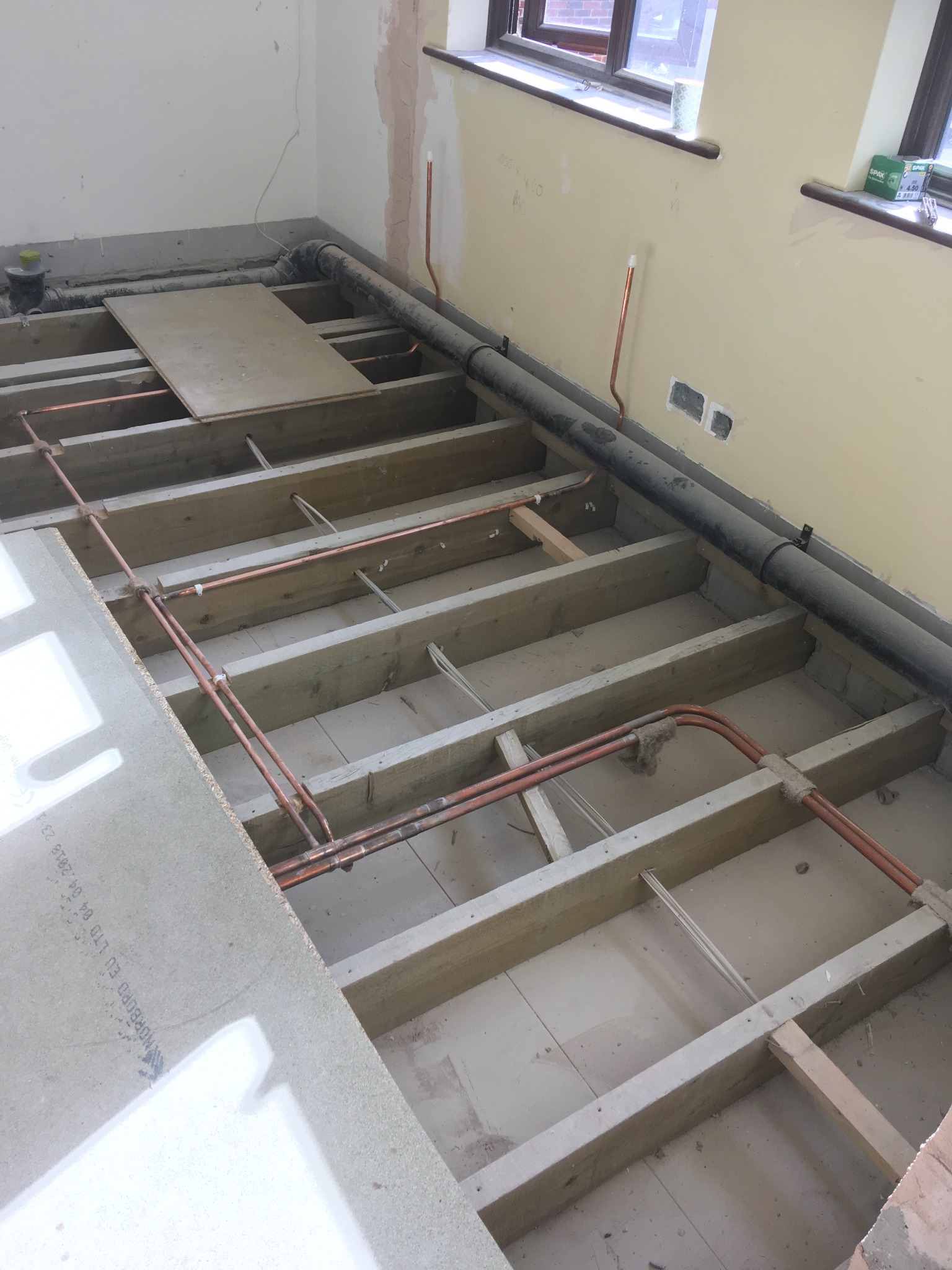 Podłoga w trakcie remontu: drewniana konstrukcja, rury miedziane, przewody elektryczne, częściowo położone płyty podłogowe, widoczne okno i ściana z instalacją elektryczną.