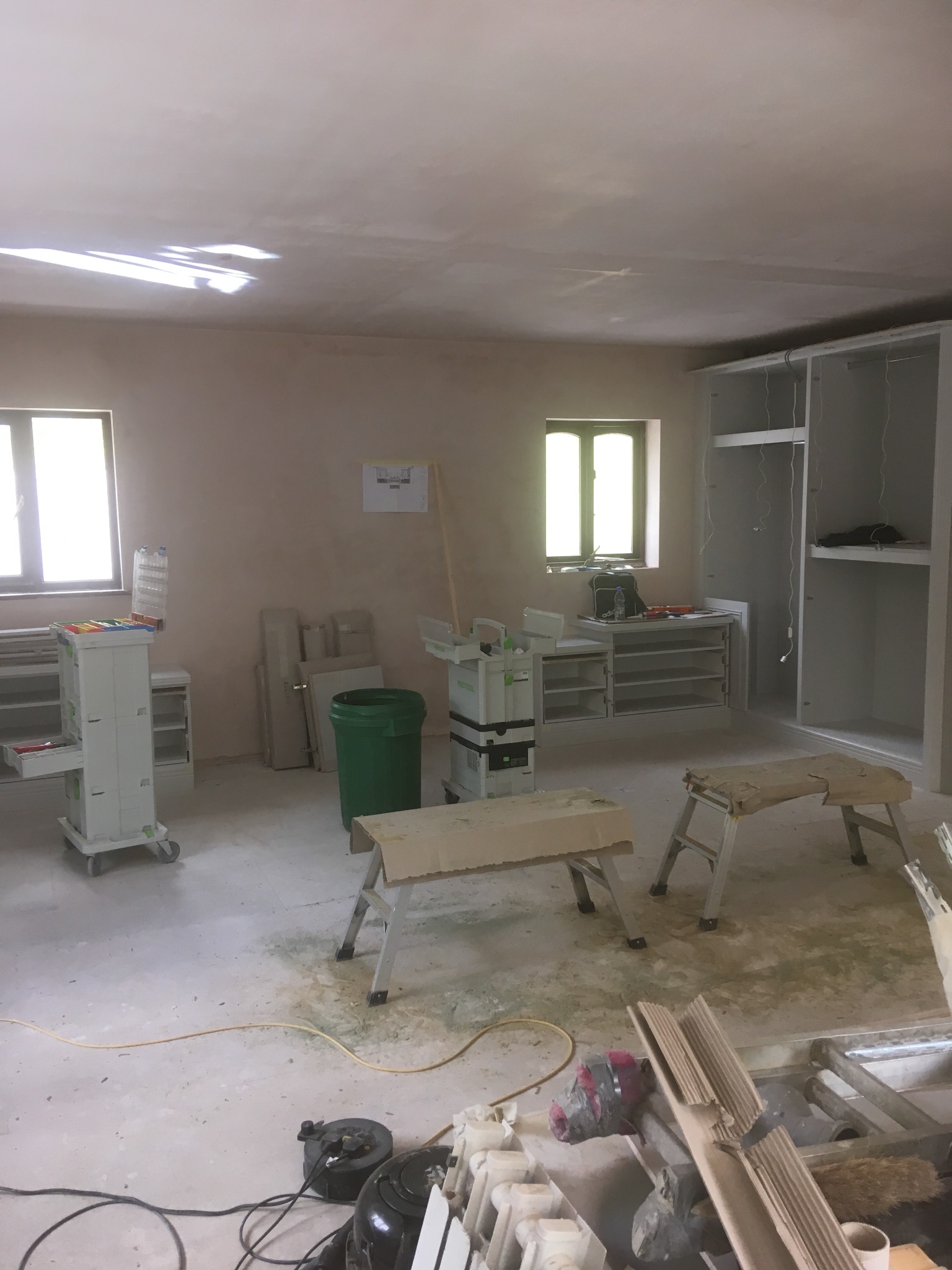 Wnętrze w trakcie montażu szafy wnękowej: widoczne narzędzia, drabiny, szafa z częściowo zamontowanymi półkami, okno z widocznymi narzędziami na parapecie, ślady po remoncie na podłodze.