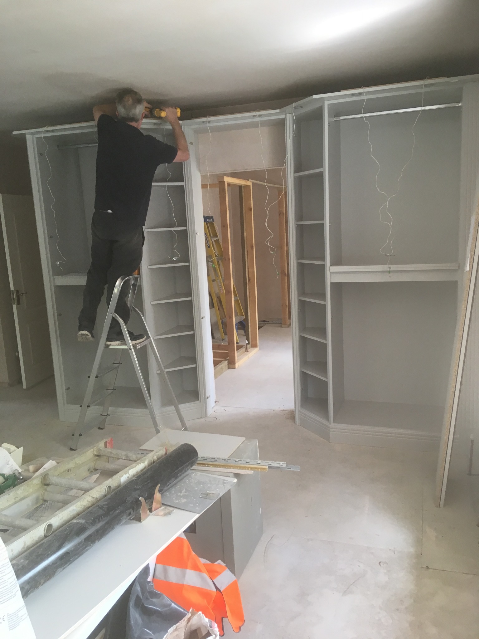 Montaż szafy wnękowej: mężczyzna na drabinie w trakcie mocowania górnej listwy szafy za pomocą wkrętarki, widoczne półki i drążki, surowy stan wykończenia wnętrza z widoczną instalacją elektryczną...