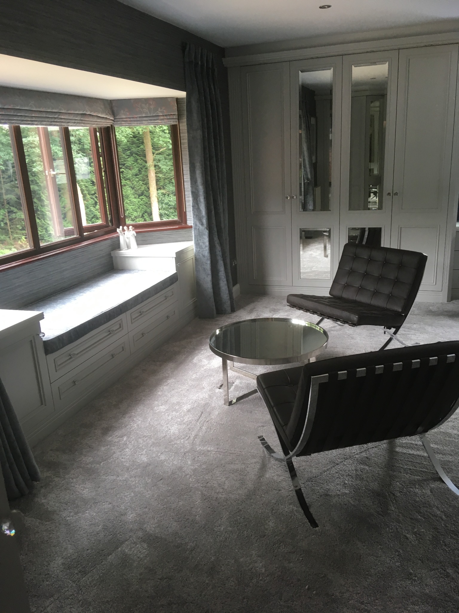 Szara szafa wnękowa z lustrami, wbudowane siedzisko przy oknie z szufladami, okrągły stolik kawowy i dwa skórzane fotele Barcelona na miękkim dywanie.