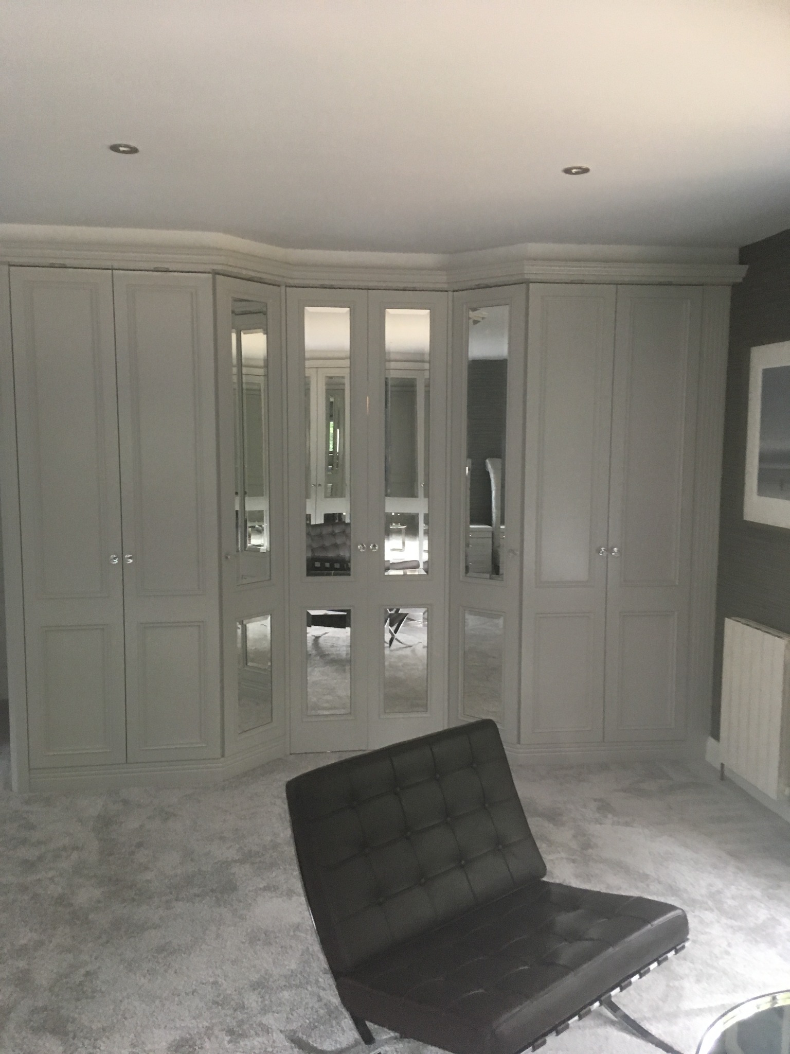 Jasnoszara szafa wnękowa z lustrzanymi drzwiami, narożna, z widocznym odbiciem wnętrza i czarnym fotelem Barcelona na tle jasnego dywanu.