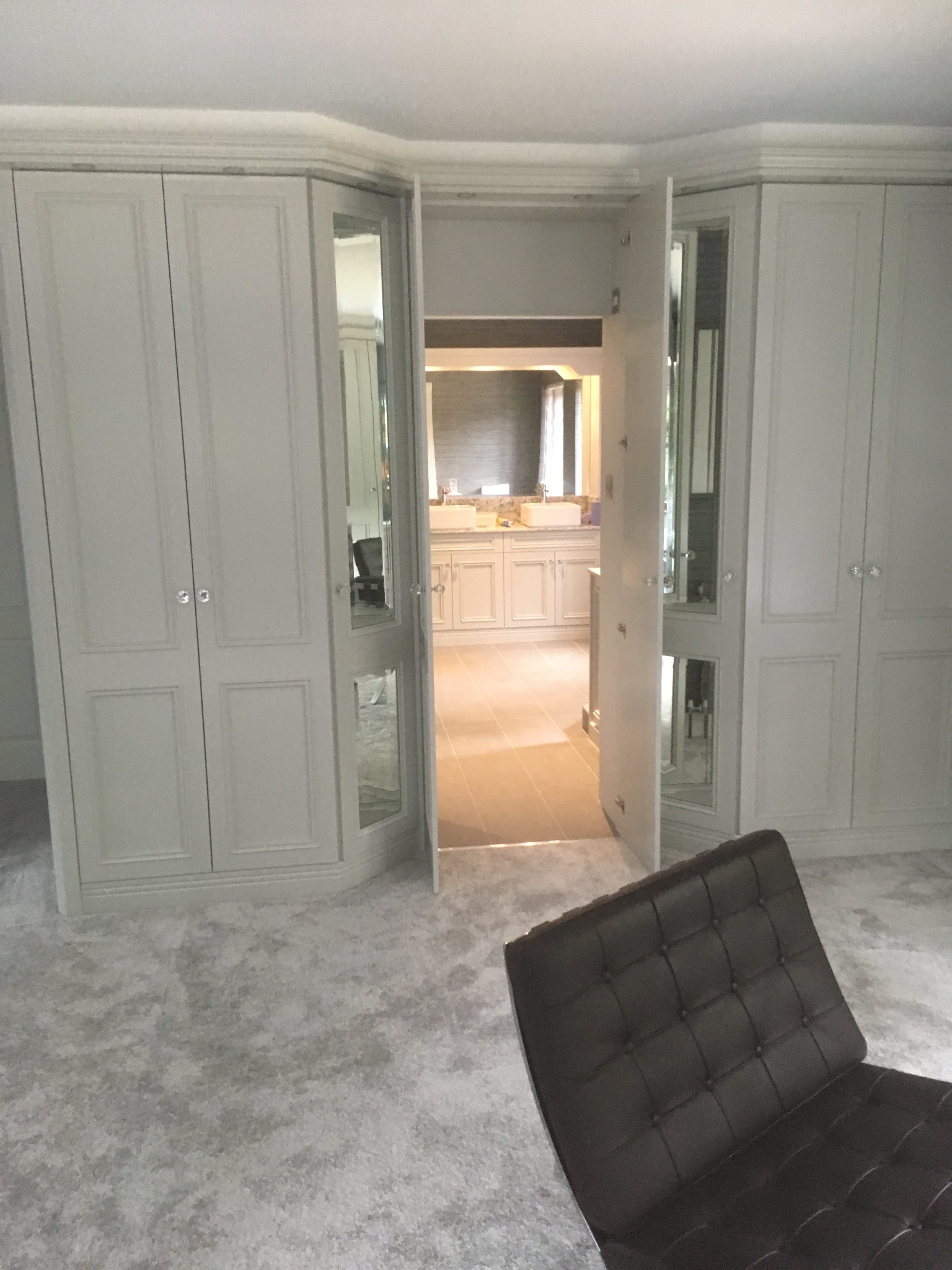 Szara szafa wnękowa z lustrami, otwarte drzwi prowadzą do łazienki z białymi szafkami, widoczny fragment brązowego fotela na pierwszym planie.