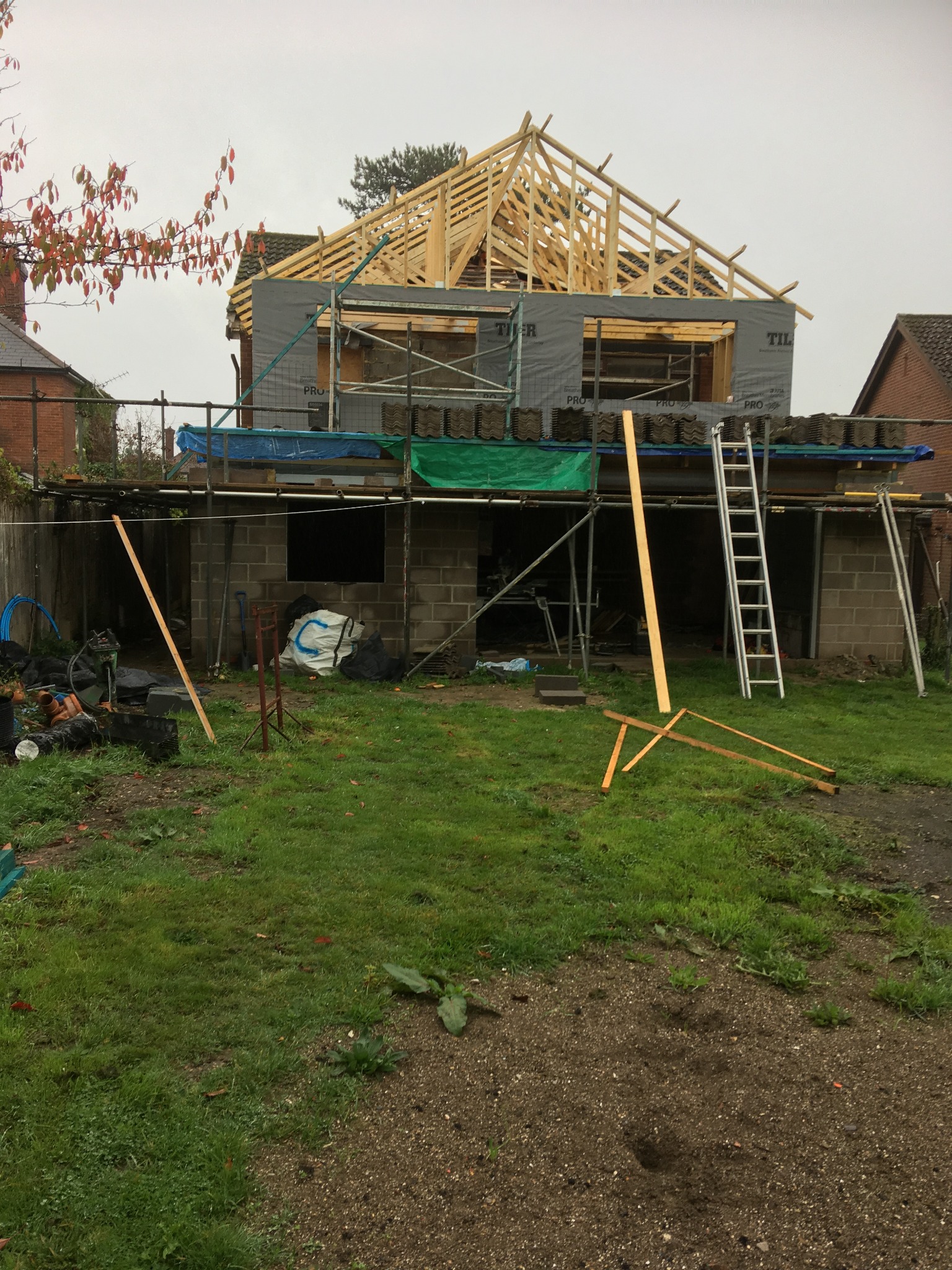 Budowa domu w trakcie realizacji: widoczne rusztowania, drewniana konstrukcja dachu, bloczki betonowe, materiały budowlane i drabina na tle zielonego trawnika.