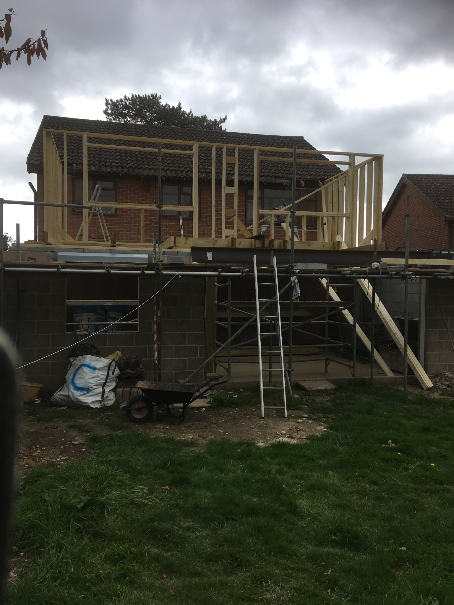 Budowa szkieletu drewnianego nad istniejącym budynkiem z cegły, widoczne rusztowania, drabiny i materiały budowlane.