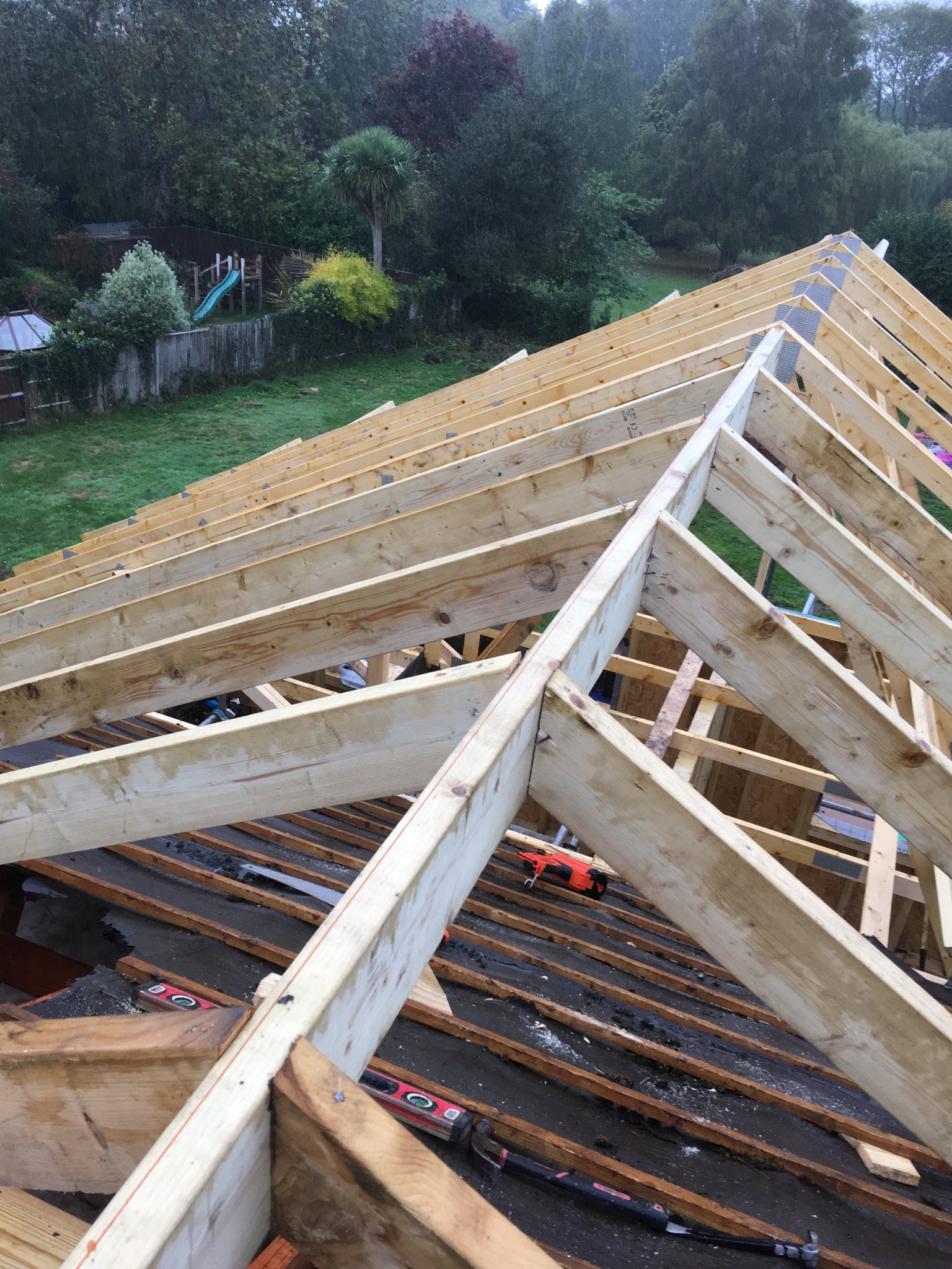 Konstrukcja drewniana dachu w trakcie budowy, widoczne krokwie, łaty, poziomice i młotek ciesielski, w tle ogród z placem zabaw.