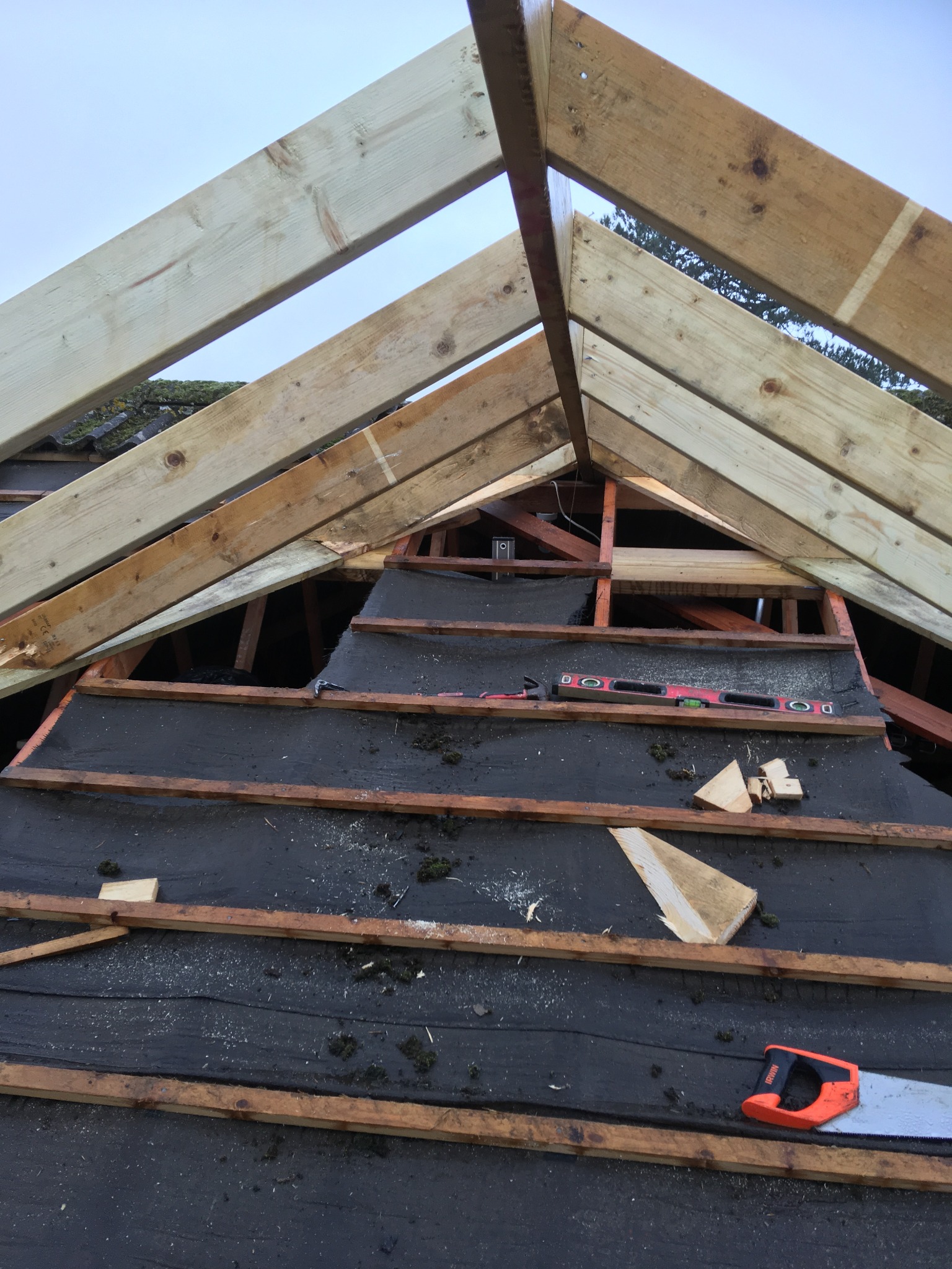 Konstrukcja drewniana dachu w trakcie budowy, widoczne krokwie, łaty, membrana dachowa, poziomica i piła ręczna.