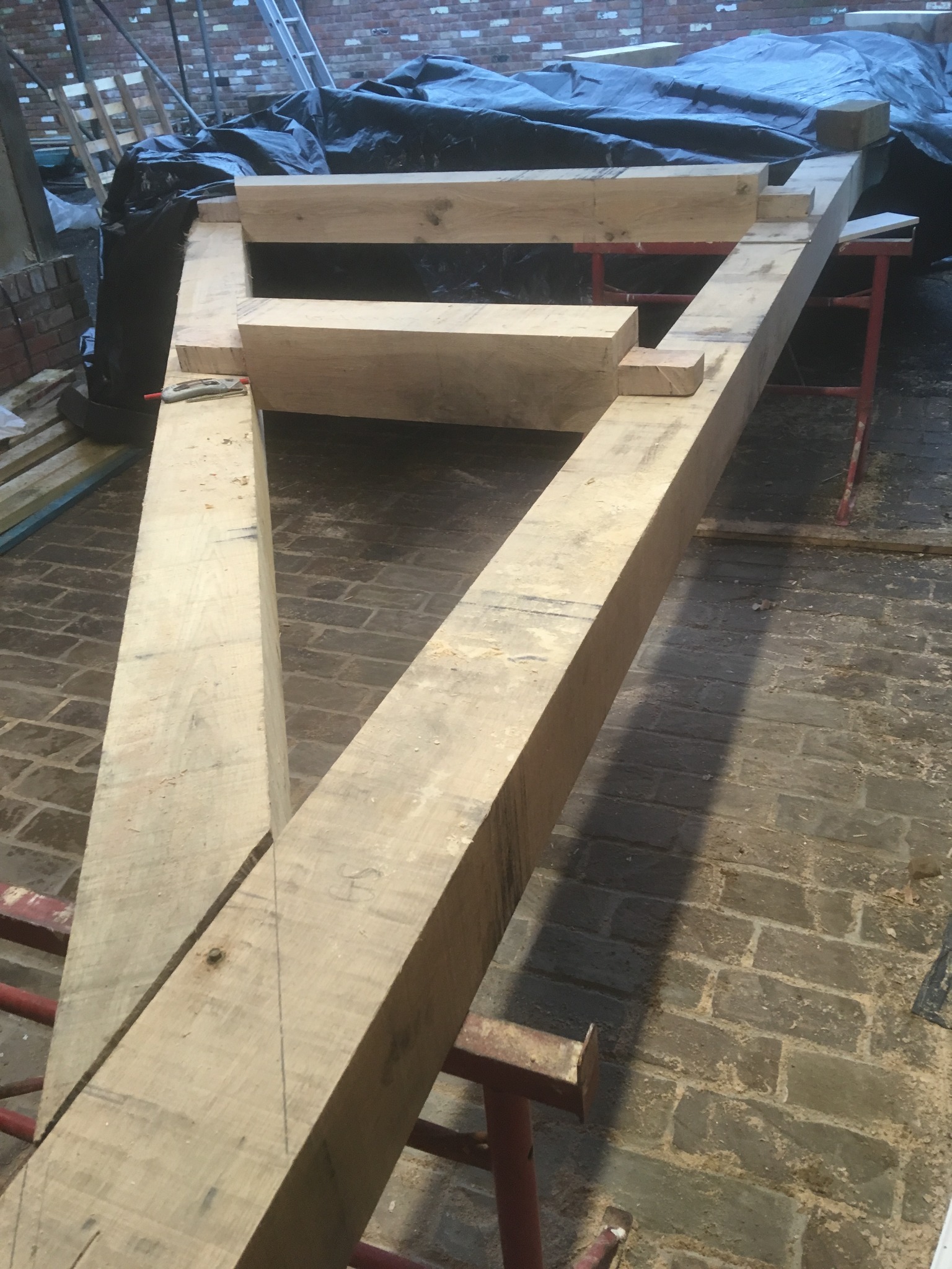 Surowe, drewniane belki tworzące konstrukcję szkieletową, częściowo podparte na czerwonych kozłach, na tle ceglanej podłogi i przykrytej plandeką przestrzeni.