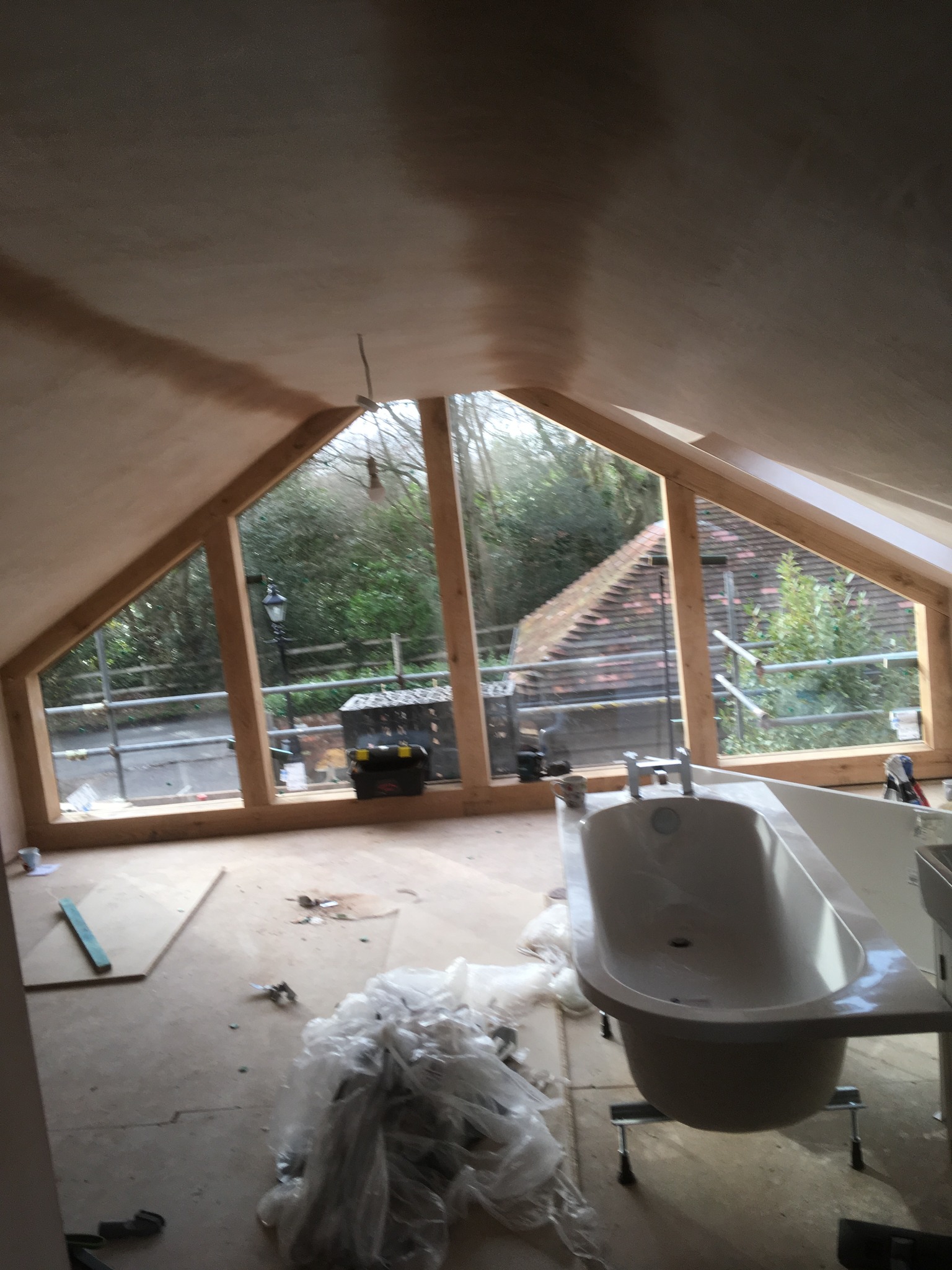 Łazienka na poddaszu w trakcie remontu: wanna wolnostojąca na metalowych nogach, okno trójkątne z widokiem na drzewa, surowy tynk na skosach dachu, narzędzia i folia ochronna na podłodze.