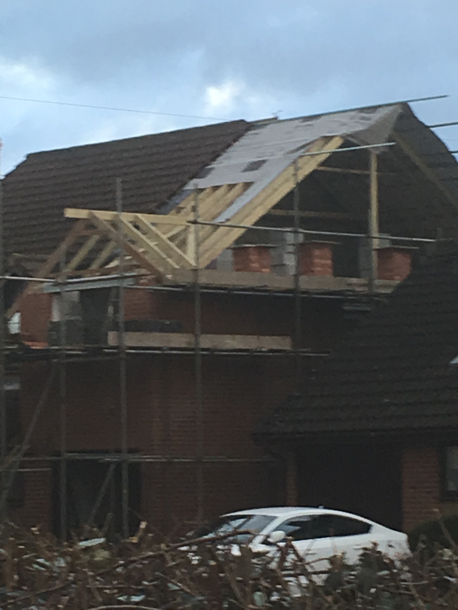 Budowa poddasza z drewnianą konstrukcją dachu i ceglanymi ścianami szczytowymi, zabezpieczona folią, widoczna z rusztowania na tle domu z brązową dachówką i białym samochodem.