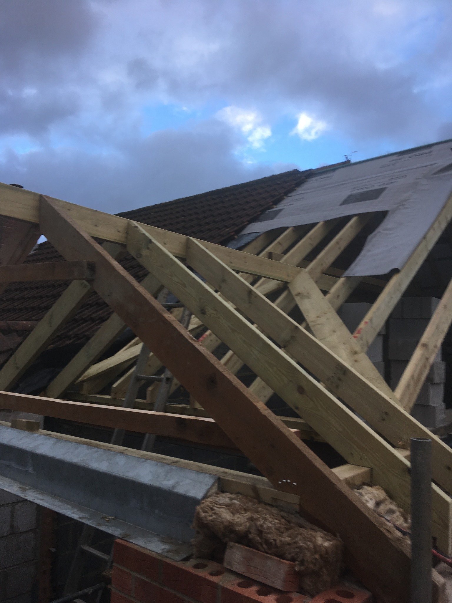 Konstrukcja drewniana dachu w trakcie budowy, widoczne krokwie, łaty, fragment pokrycia dachowego i izolacja, pochmurne niebo w tle.