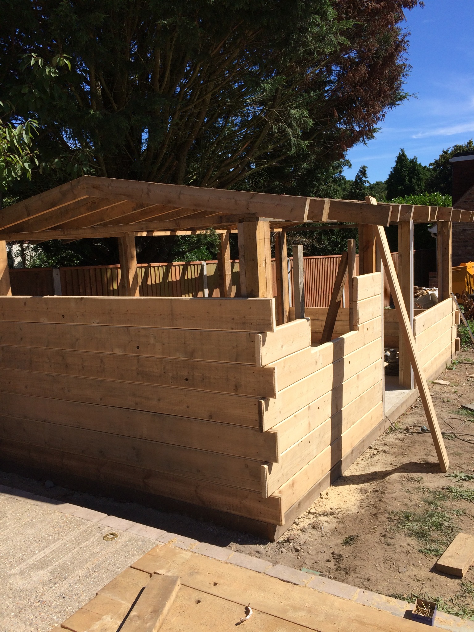 Konstrukcja z jasnego drewna w trakcie budowy, prawdopodobnie altana lub domek narzędziowy, z widocznymi elementami szkieletu i deskami tworzącymi ściany, na tle zieleni i błękitnego nieba.