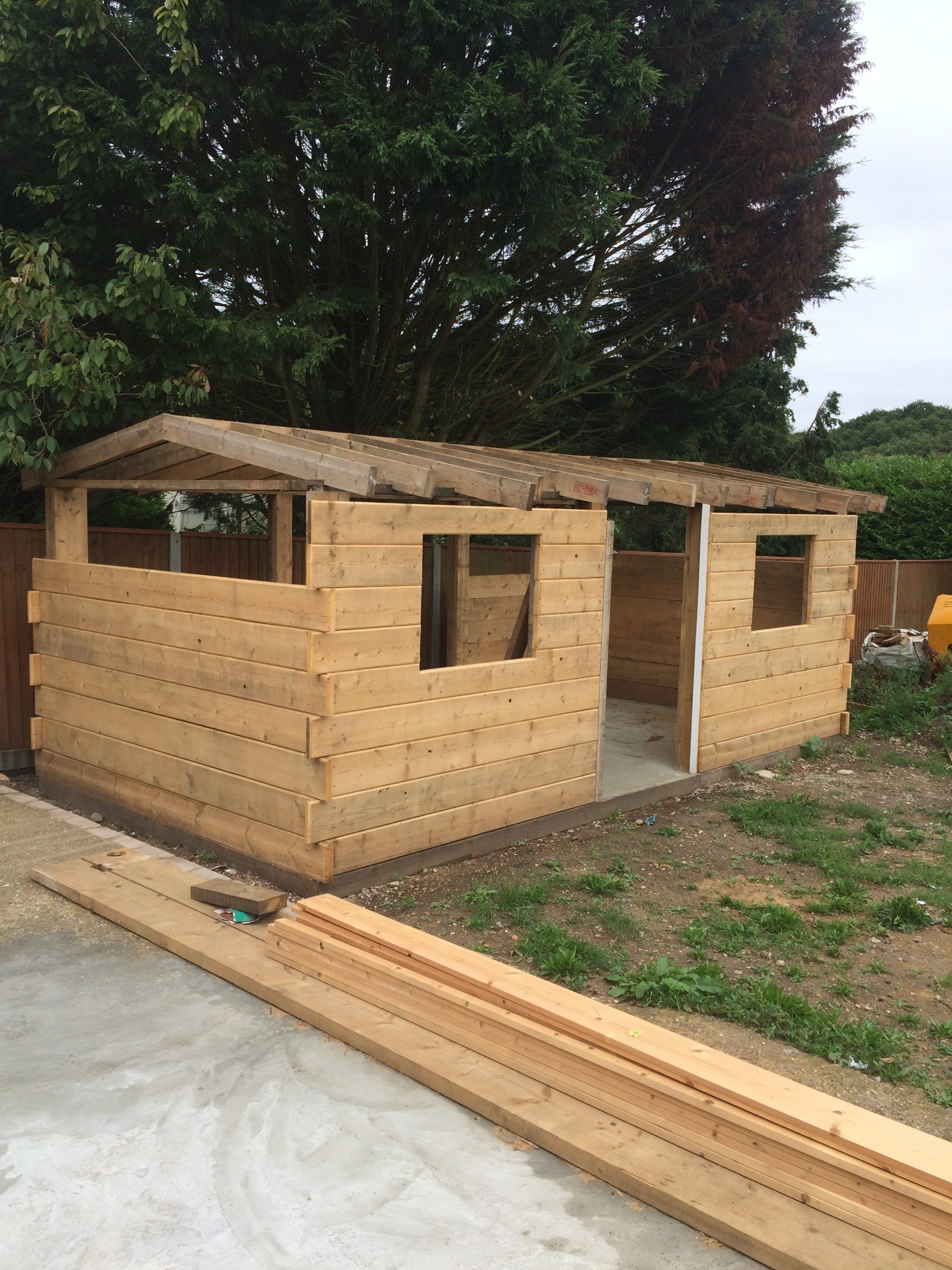 Surowa konstrukcja drewnianej szopy ogrodowej z widocznymi elementami dachu, ścianami z desek i stertą drewna na betonowej płycie przed budowlą, na tle zieleni.