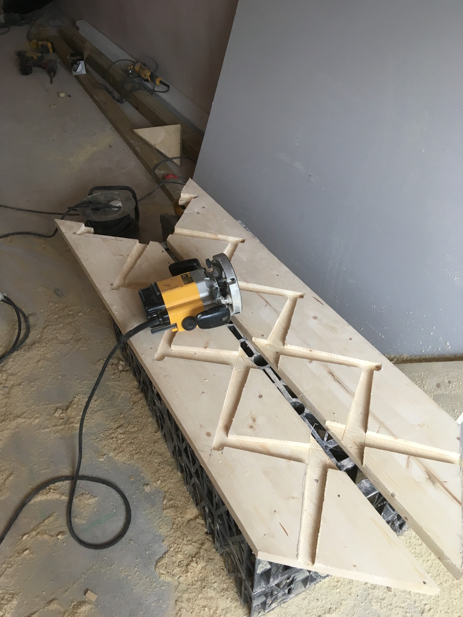 Frezarka na drewnianej płycie z wyfrezowanymi geometrycznymi wzorami w warsztacie, widoczne wióry i inne narzędzia w tle.