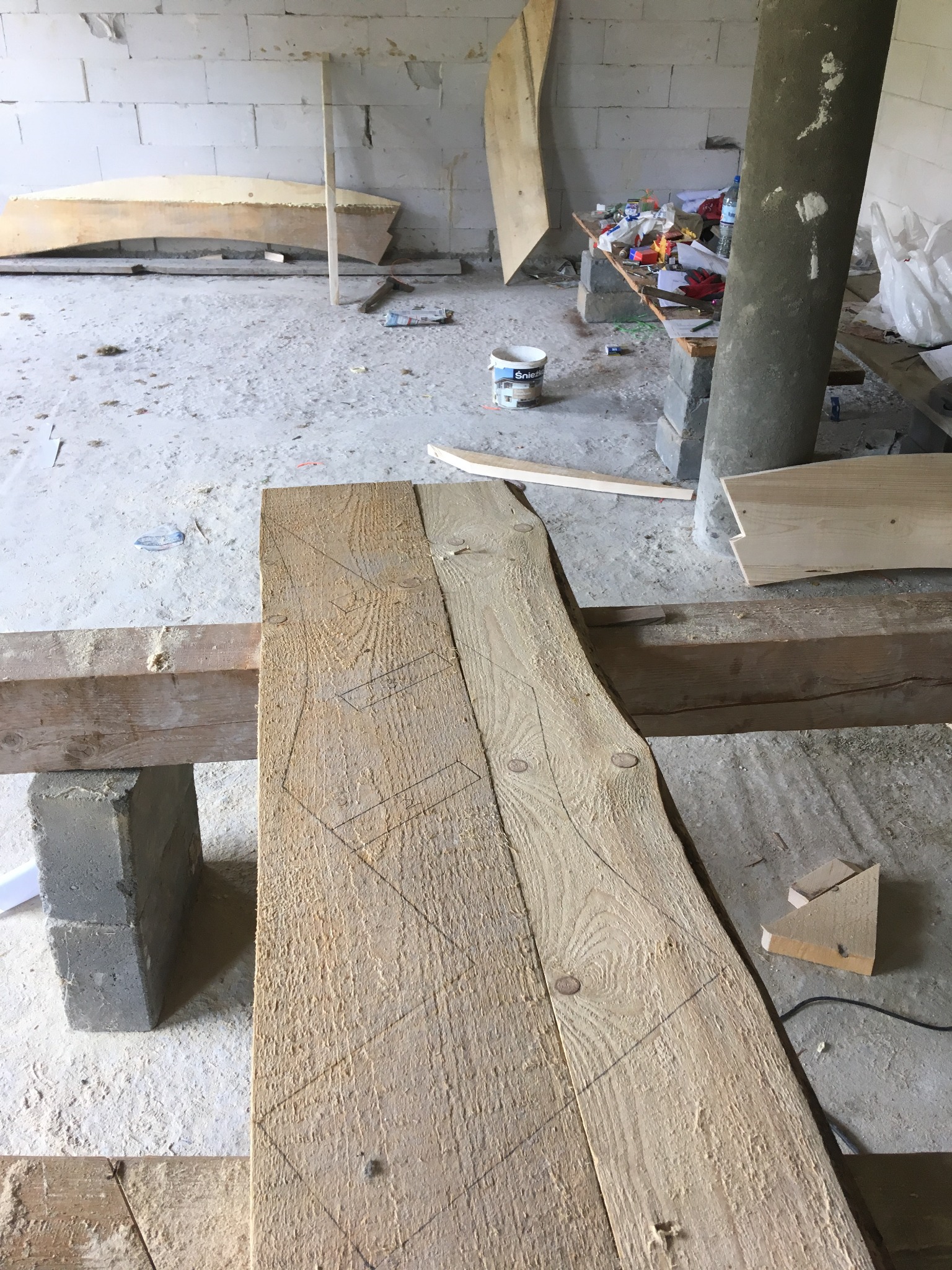 Drewniana deska z naniesionymi liniami cięcia, wsparta na betonowym bloczku w surowym wnętrzu, widoczne narzędzia i materiały budowlane w tle.