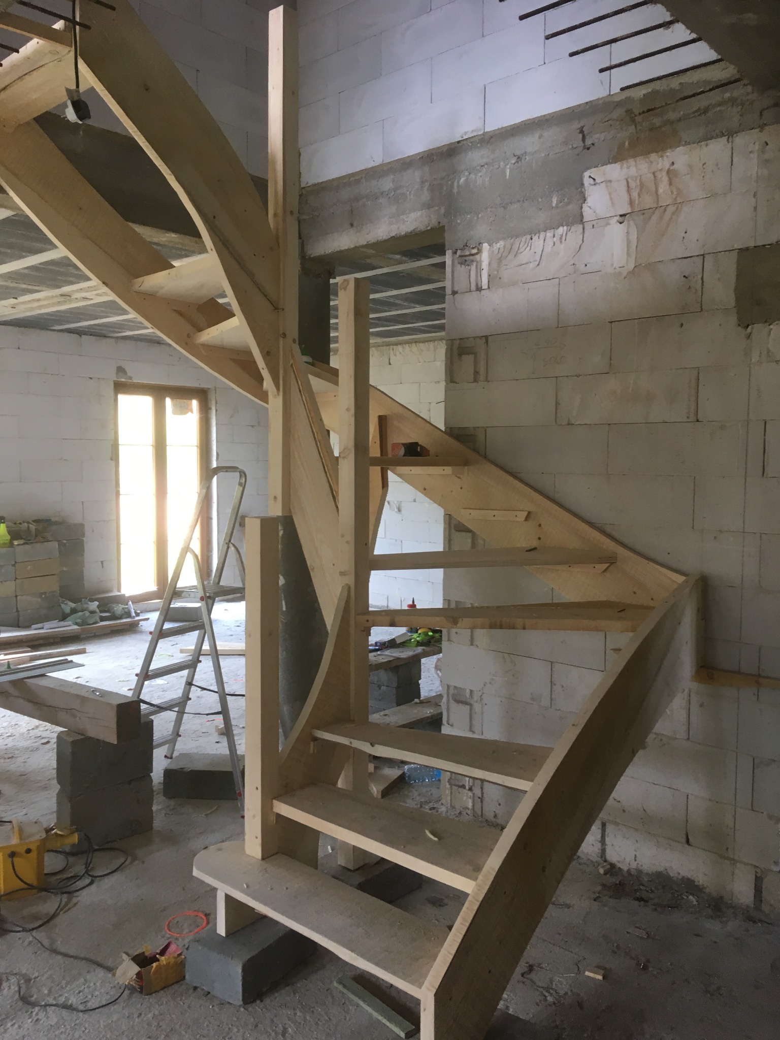 Surowe drewniane schody spiralne w trakcie montażu w surowym wnętrzu z białych bloczków, widoczne narzędzia i drabina.