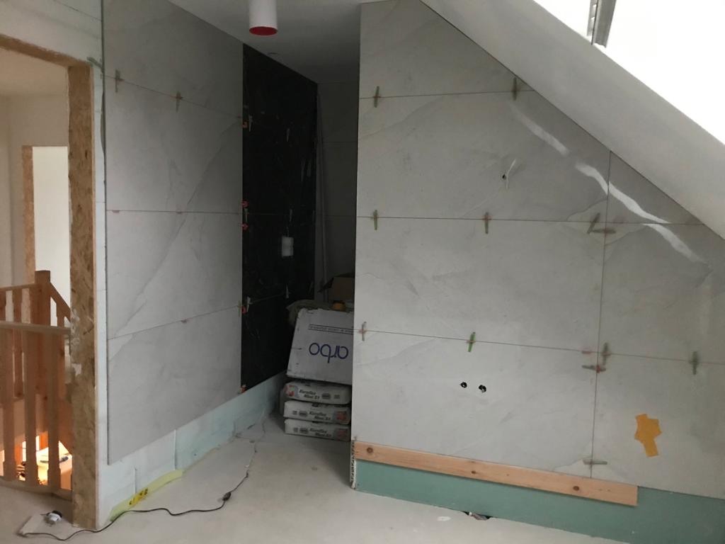 Ściana łazienki w trakcie wykańczania: jasne płytki imitujące marmur układane na skosie poddasza, widoczne krzyżyki dystansowe, poziomica i worki z zaprawą.