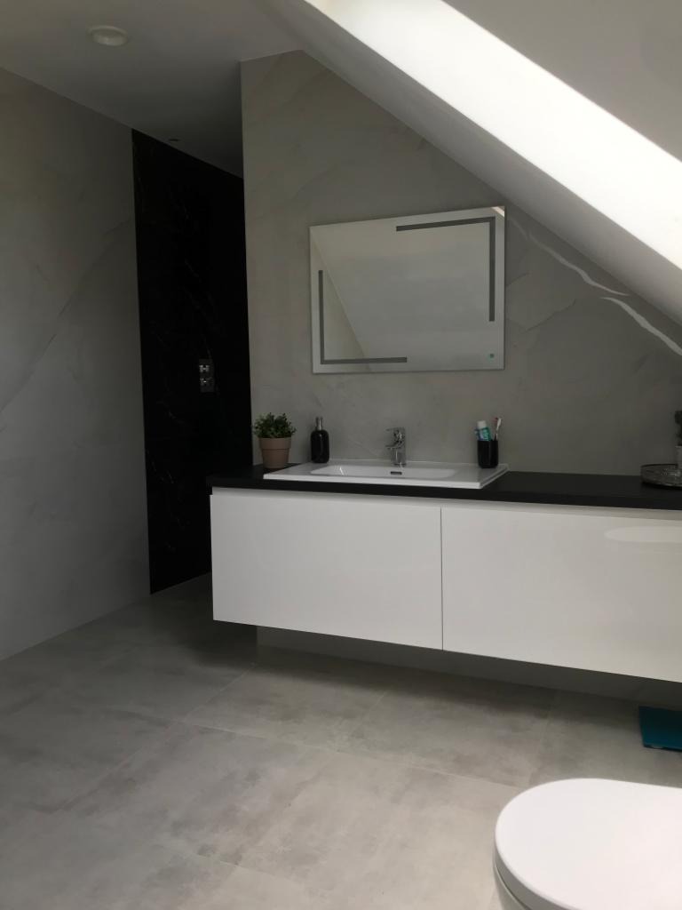 Nowoczesna łazienka na poddaszu z białą szafką pod umywalką, czarnym blatem, podświetlanym lustrem i czarną kabiną prysznicową.