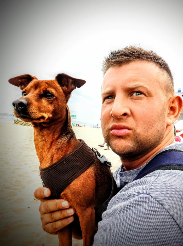 Mężczyzna z zadartą brwią trzyma na rękach brązowego pinczera w brązowej uprzęży na piaszczystej plaży z widocznymi ludźmi w tle.