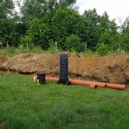 Prace ziemne: Wykopany rów z widocznymi rurami kanalizacyjnymi i studzienką, mała koparka i niwelator laserowy w tle, zielona trawa.