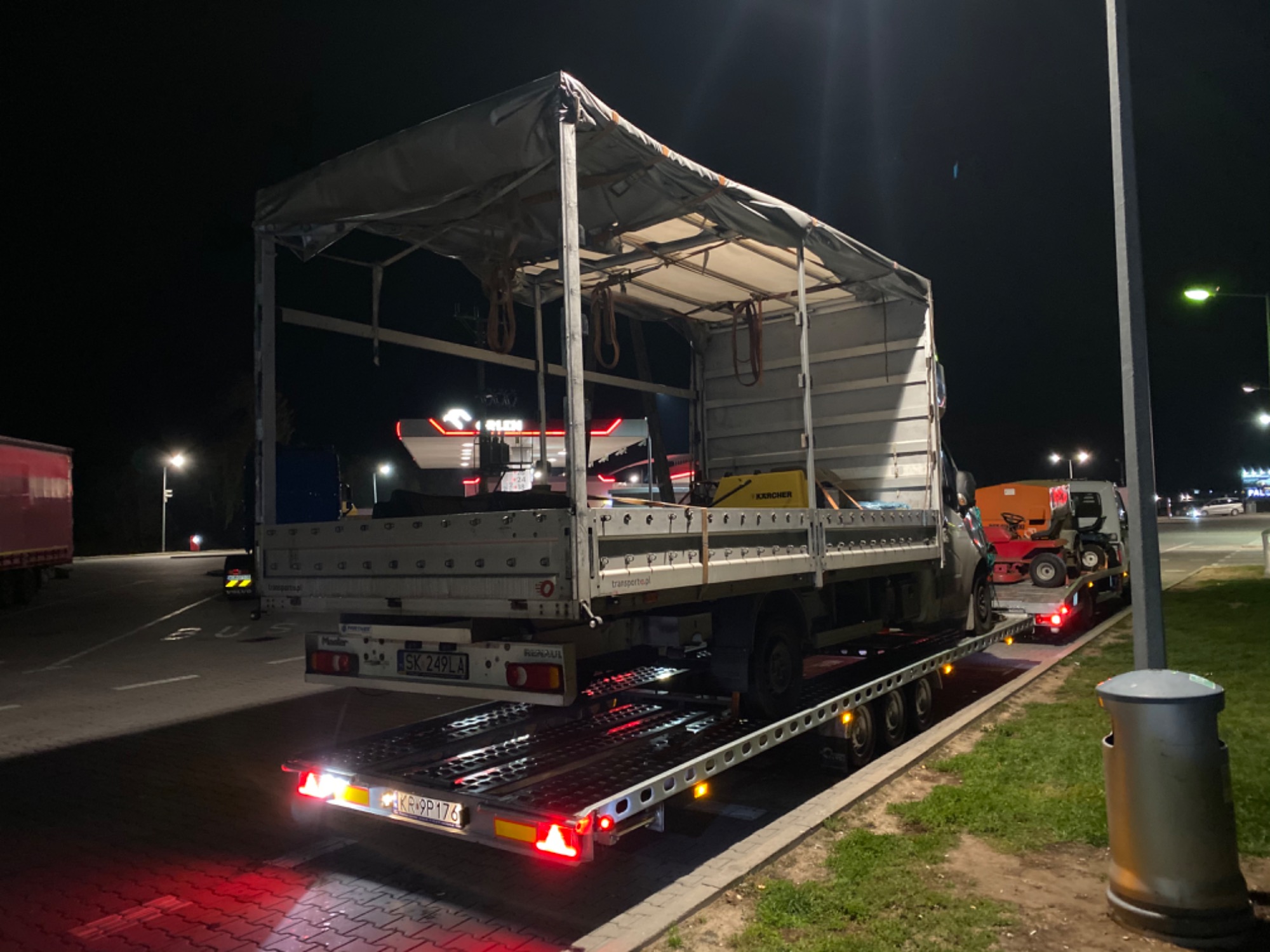 Nocny transport samochodem dostawczym z plandeką, przewożącym myjkę ciśnieniową Karcher i kosiarkę traktorek, na platformie z oświetleniem, widok z tyłu na parkingu z oświetleniem ulicznym.
