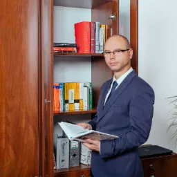 Mężczyzna w garniturze i okularach stoi na tle regału z książkami prawniczymi, trzymając otwartą książkę, z widocznym tytułem 'Postępowanie Karne' na jednej z półek.