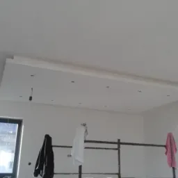 Wnętrze w trakcie remontu: widok na sufit z zamontowaną płytą kartonowo-gipsową, rusztowanie robocze z ubraniami i narzędziami, surowa żarówka zwisająca z sufitu.