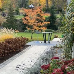 Ogród z ścieżką z jasnoszarej kostki granitowej, obrzeża z lawendy i hortensji bukietowych w odcieniach czerwieni, trawnik z drzewami iglastymi i klonem o pomarańczowych liściach, w tle mała...