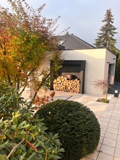 Nowoczesny dom z garażem, w którym składowane jest drewno na opał, otoczony zadbaną zielenią: krzewami, drzewami liściastymi w jesiennych barwach i iglakiem. Ścieżka z kostki prowadzi do wejścia.